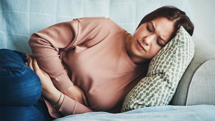 A dark-haired woman is lying on a sofa and holding her tummy as she winces in pain
